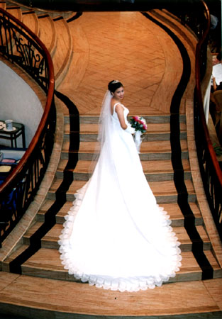Bride in extremely long, flowing white gown holding flowers and ascending a wooden staircase